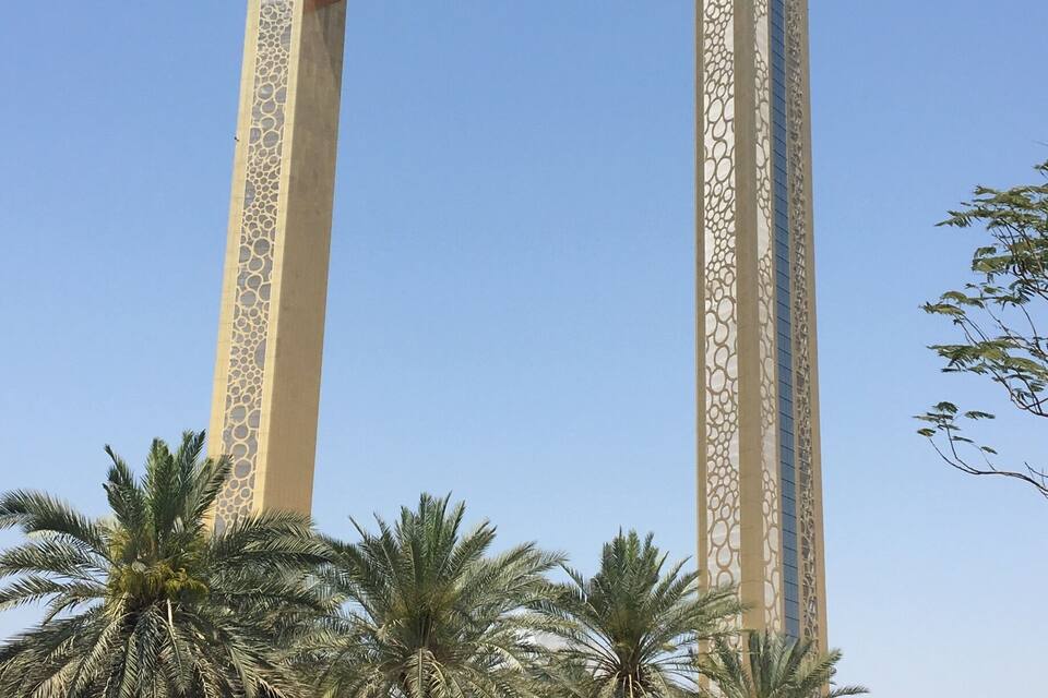 Um arco alto e ornamentado, com padrões elaborados, cercado por palmeiras e um gramado bem cuidado.
