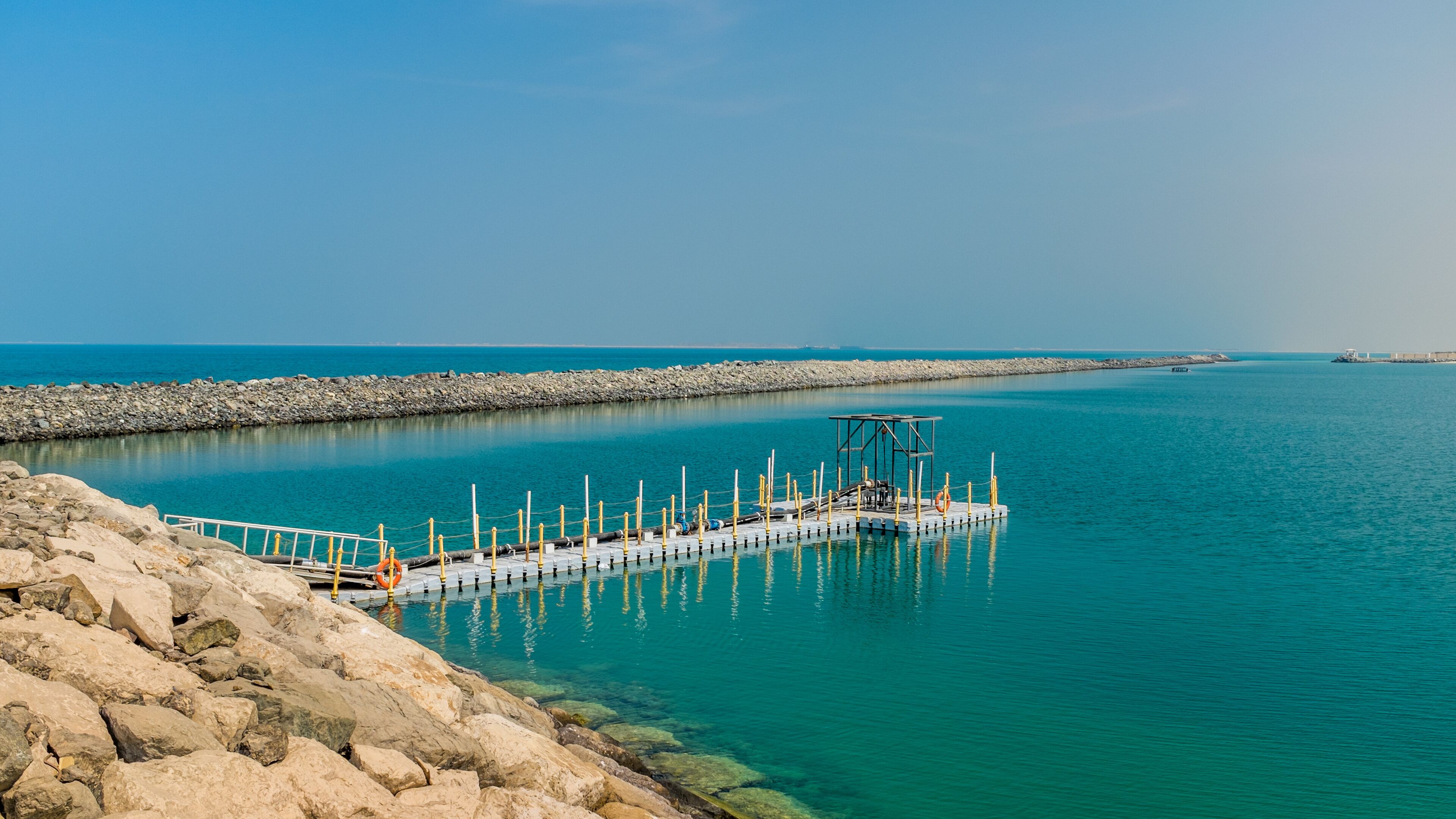 A pier extending into a calm turquoise sea, surrounded by rocky shores and a clear sky.