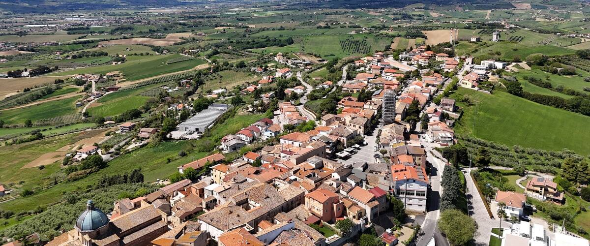 A town with terracotta-roofed buildings, surrounded by green fields and mountains in the background.