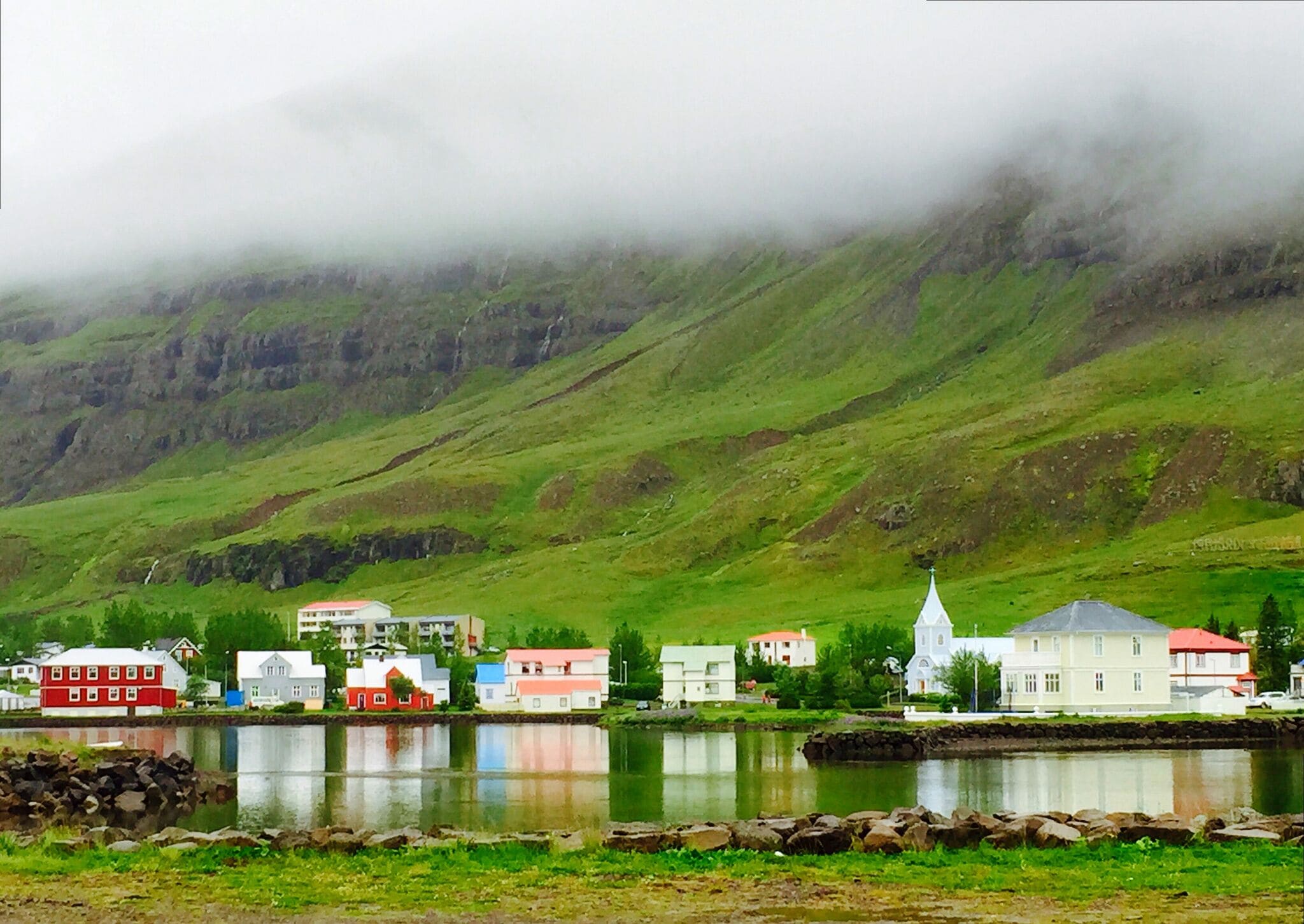 A coastal village with colorful houses and a church by a calm lake, surrounded by green hills and mist.