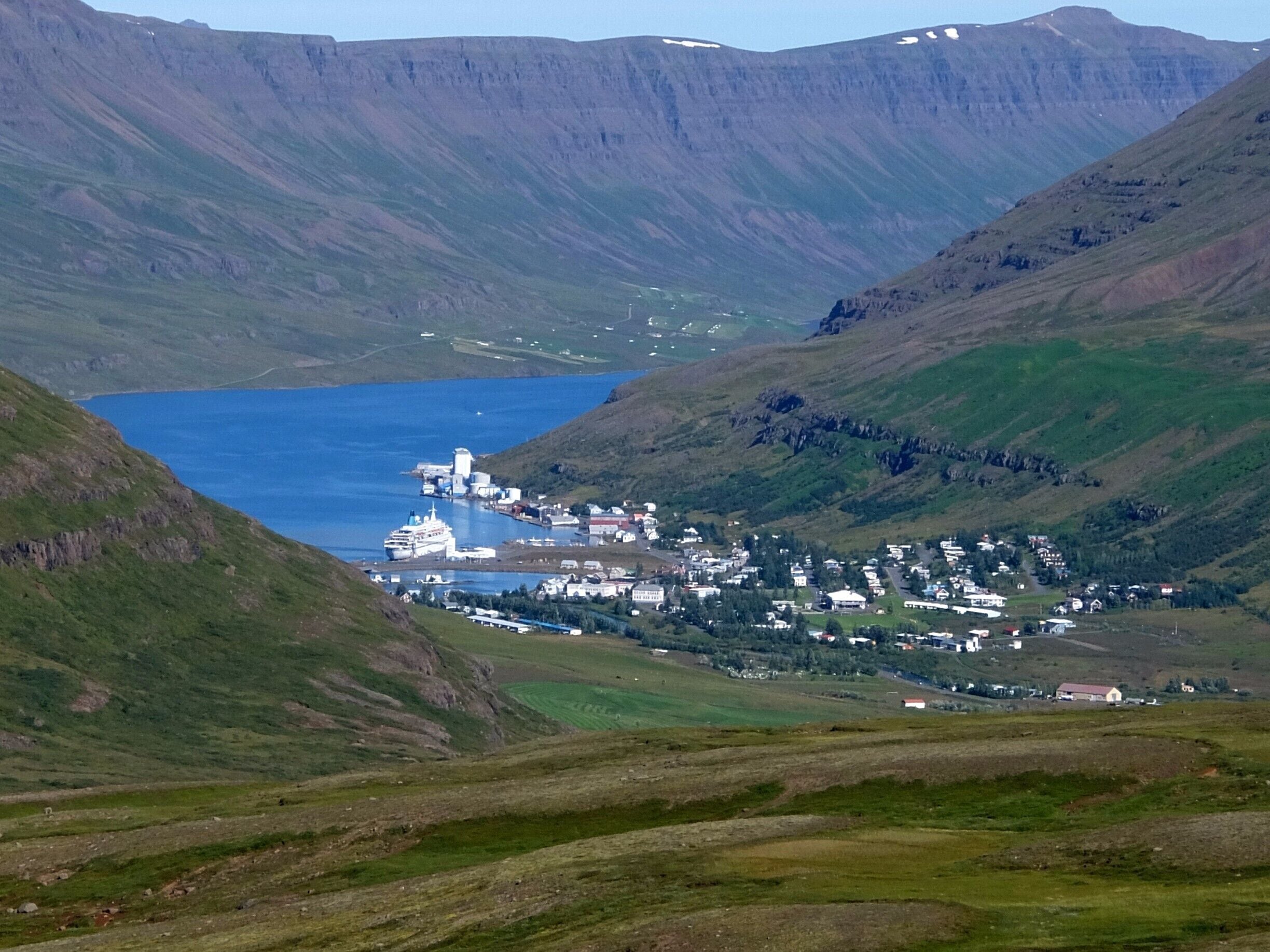 A coastal town with a harbor, surrounded by green hills and a large body of water.