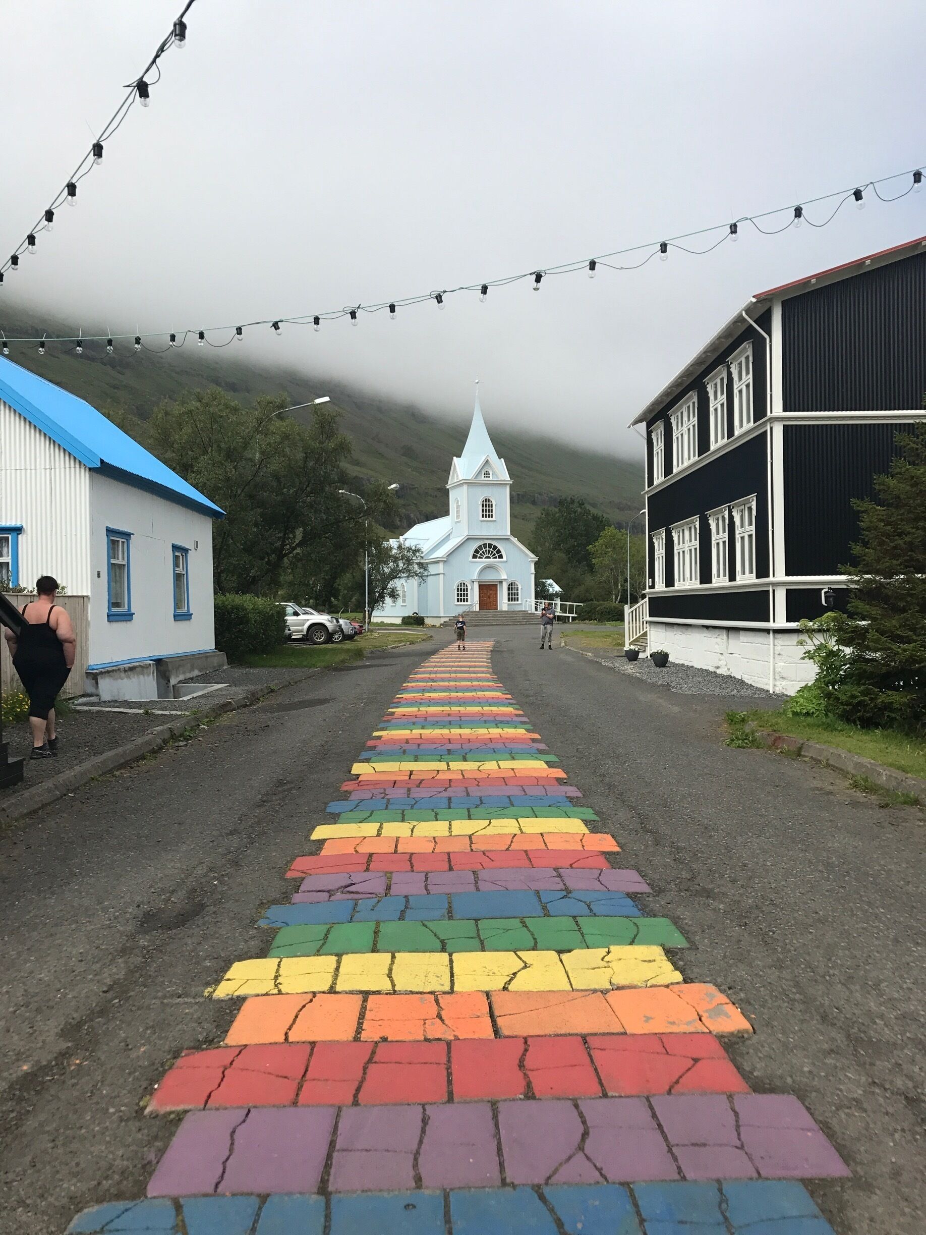 A rainbow-colored pedestrian path leading to a white church with a steeple, surrounded by buildings and a mountainous backdrop.
