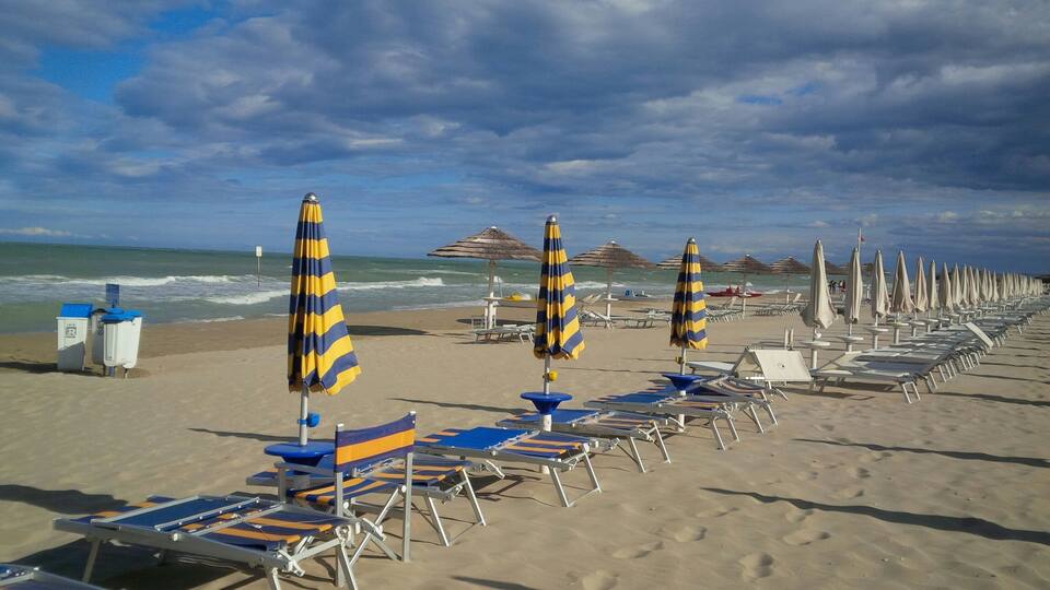 A beach with rows of sun loungers and striped umbrellas, a few beach huts, and the ocean in the background.