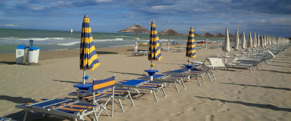 A beach with rows of sun loungers and striped umbrellas, a few beach huts, and the ocean in the background.