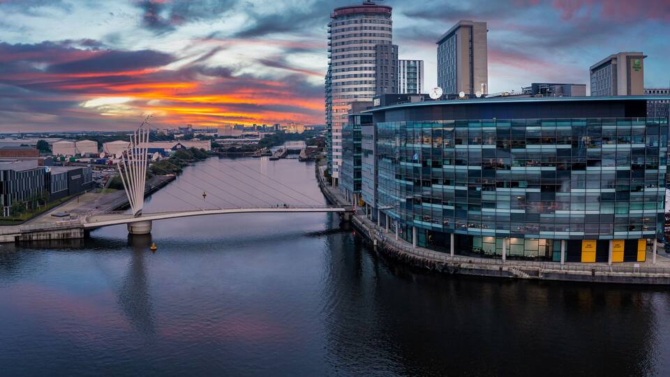 A river with a bridge, modern buildings, and a sunset sky.