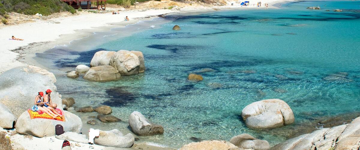 A coastal landscape with clear turquoise waters, large rocks, and a sandy beach.