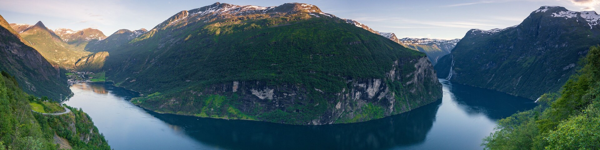 A fjord surrounded by steep mountains with patches of snow, a winding road, and a calm body of water.
