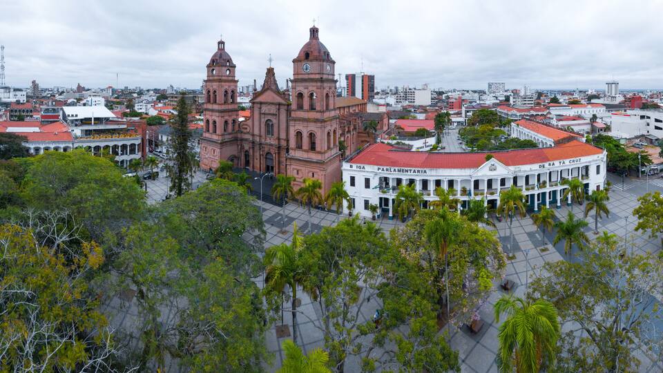 A historic church with two towers, surrounded by a plaza with palm trees and a white building with a red roof.