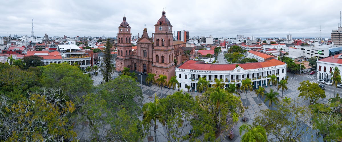 A historic church with two towers, surrounded by a plaza with palm trees and a white building with a red roof.