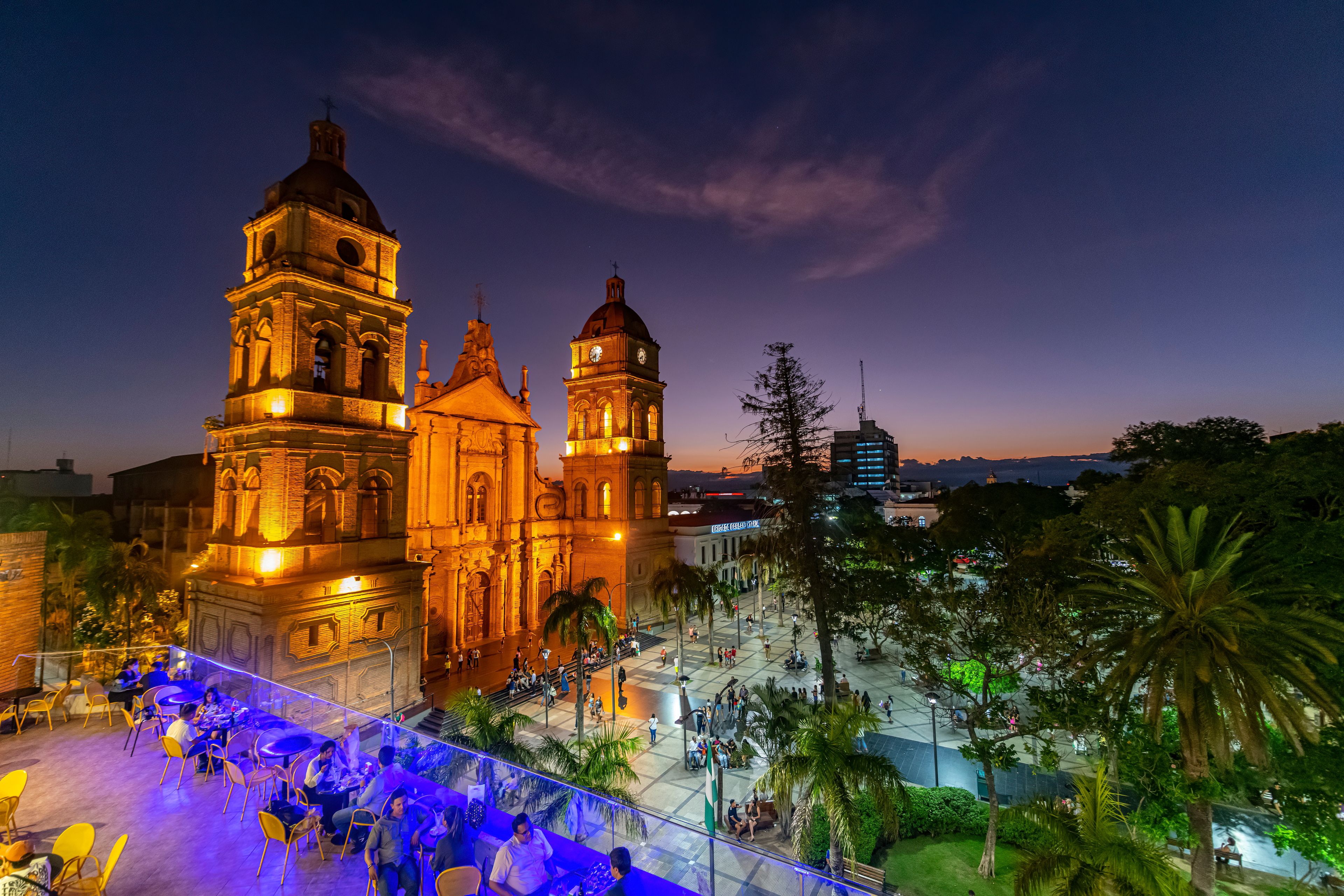 Una iglesia histórica con dos torres destacadas, una plaza con gente y un atardecer de fondo.