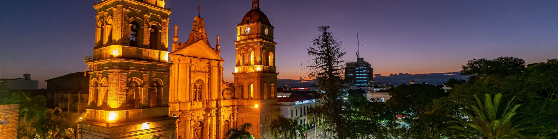 Una iglesia histórica con dos torres destacadas, una plaza con gente y un atardecer de fondo.