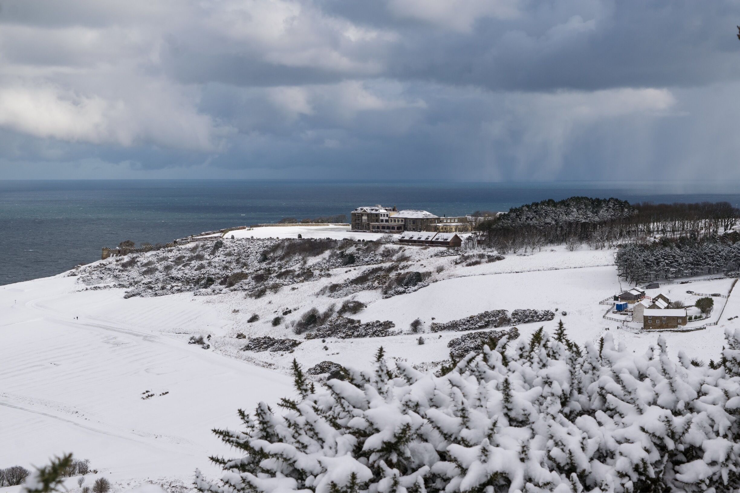 A snowy landscape with a large building and a smaller structure near a body of water.
