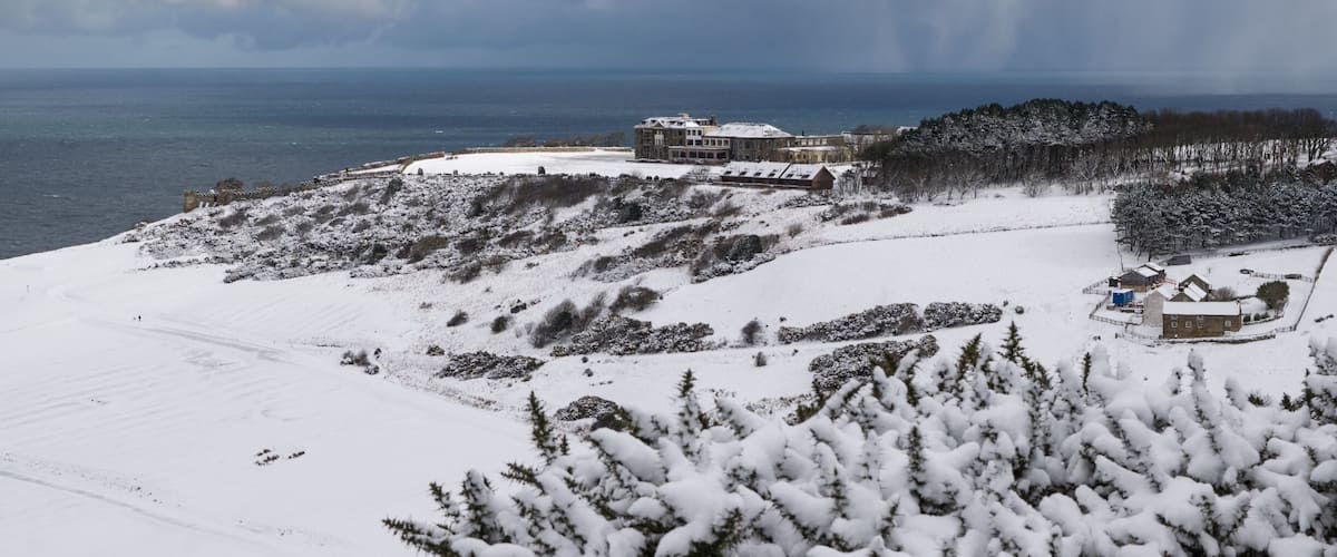 A snowy landscape with a large building and a smaller structure near a body of water.