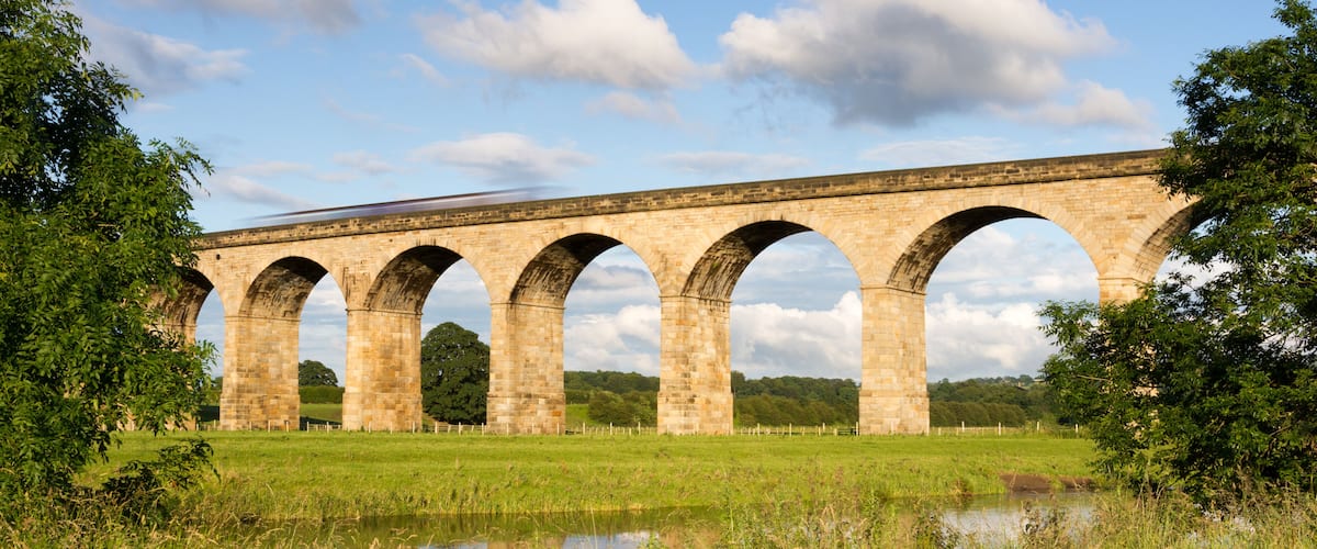 A stone viaduct with multiple arches spanning a river, surrounded by green fields and trees.