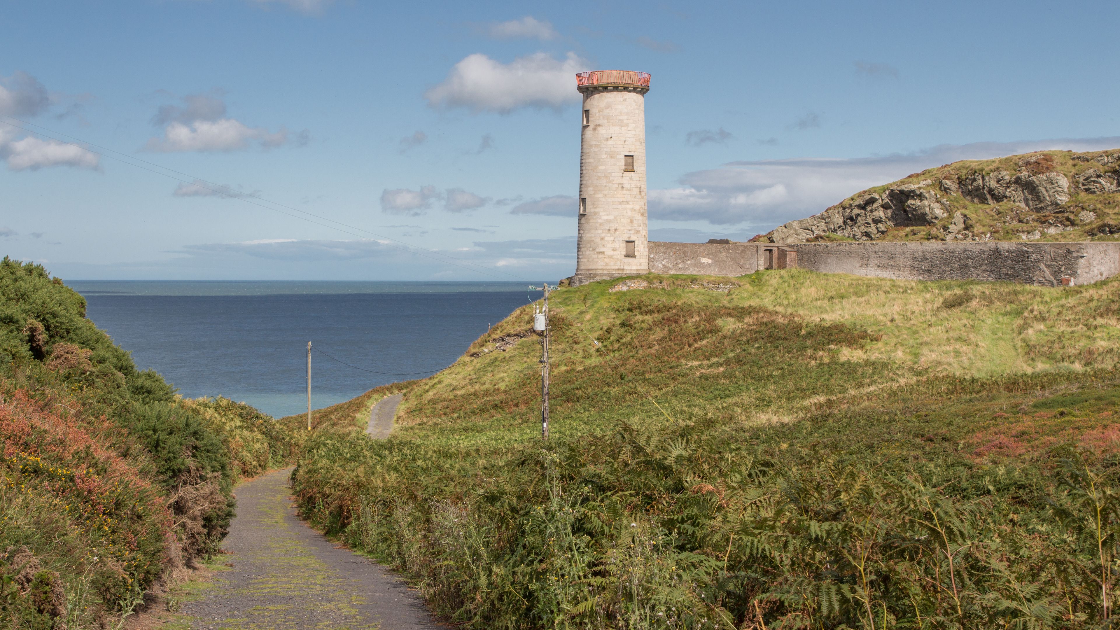 Een kustpad leidt naar een stenen vuurtoren met een rode top, omgeven door grasland en een rotsachtige klif.