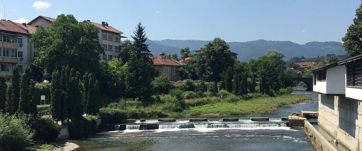 A river with a small waterfall, surrounded by buildings and trees.