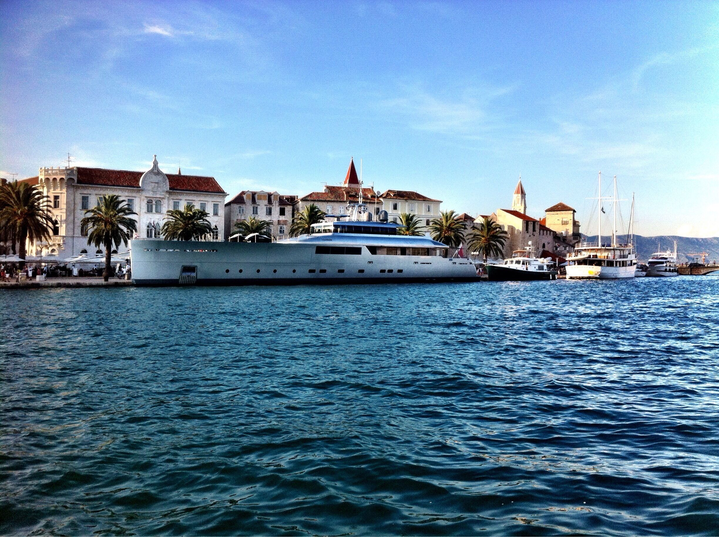 A large yacht docked at a harbor with historic buildings and palm trees in the background.