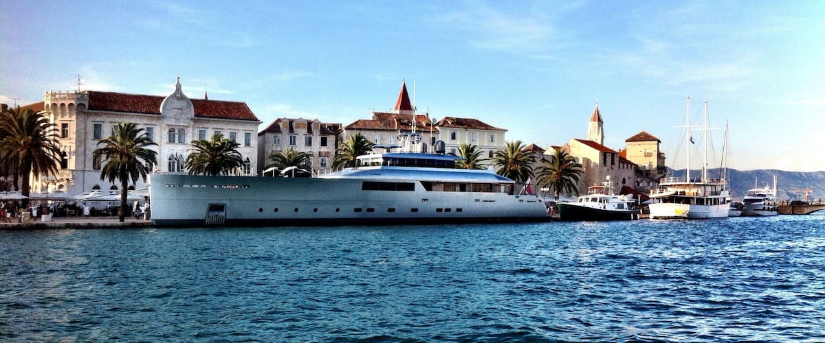 A large yacht docked at a harbor with historic buildings and palm trees in the background.