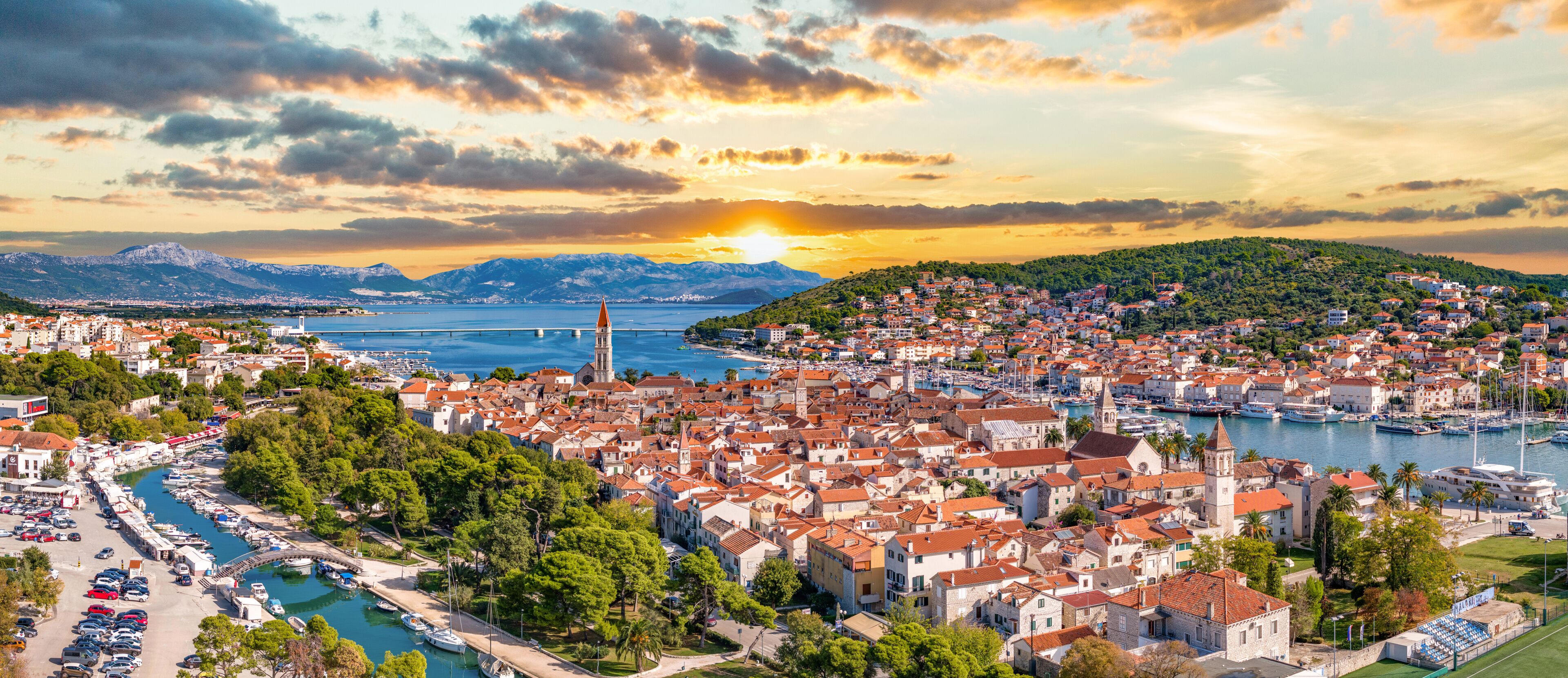 A coastal cityscape with a marina, a prominent church, and a mountain range in the background.