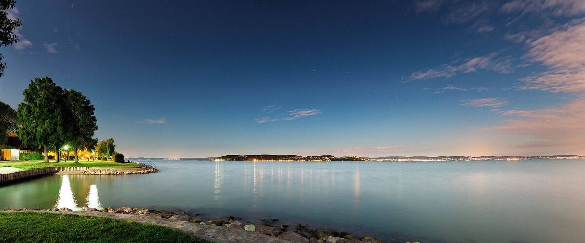 A calm lake at dusk with a distant shoreline and trees on the left.