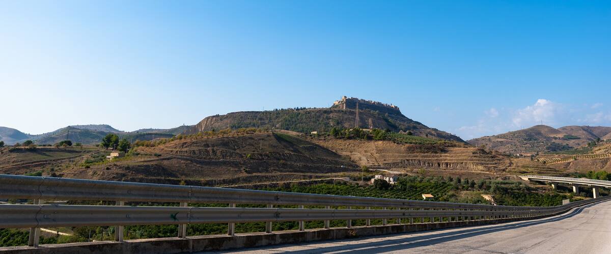 A highway with a guardrail, a hill with a castle-like structure, and a bridge in the background.