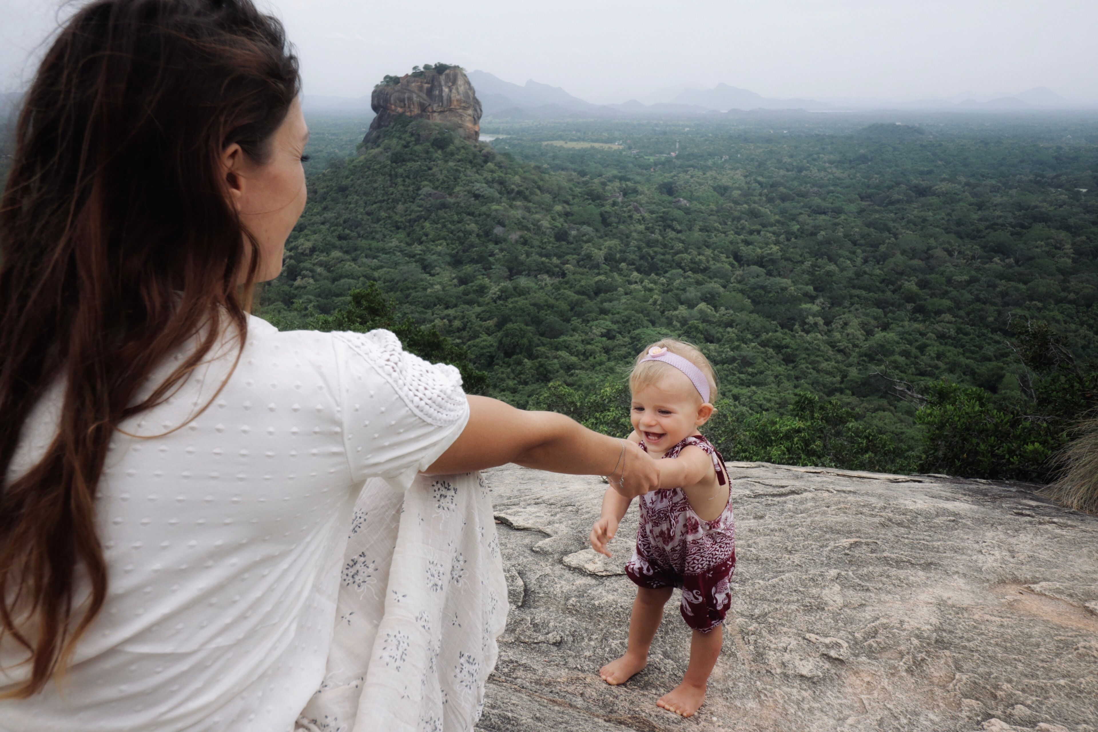 Una mujer y un niño en un afloramiento rocoso, con vista a un bosque denso y una formación rocosa destacada.