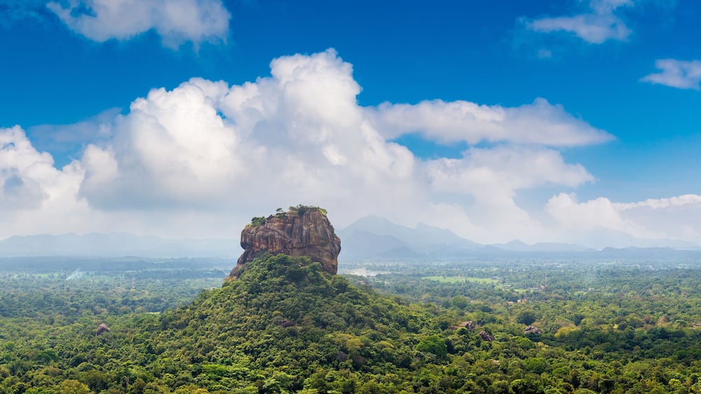 A prominent rock formation surrounded by dense green forest and a vast landscape under a blue sky with scattered clouds.