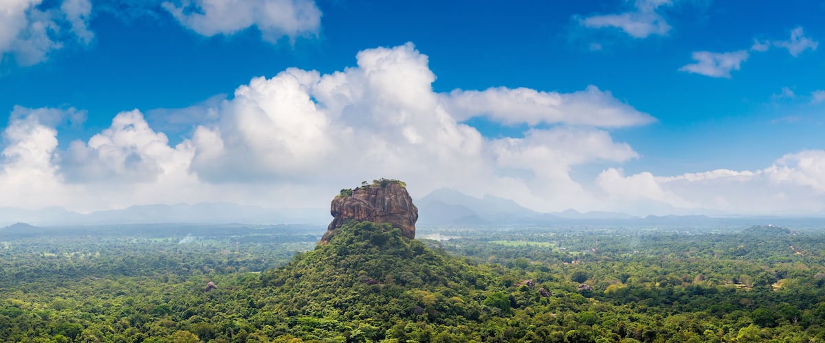 Una imponente formación rocosa rodeada de un bosque verde frondoso y un extenso paisaje bajo un cielo azul con nubes dispersas.