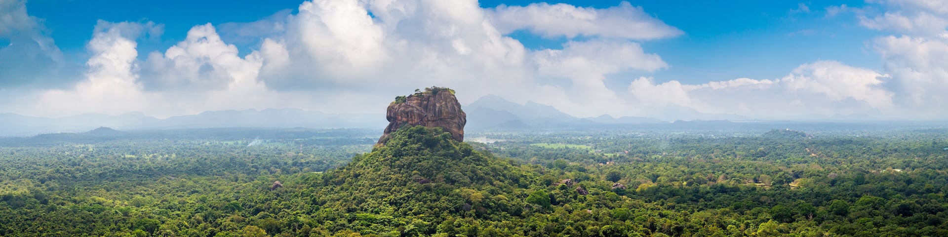 A prominent rock formation surrounded by dense green forest and a vast landscape under a blue sky with scattered clouds.