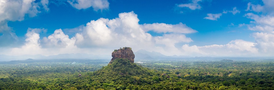 A prominent rock formation surrounded by dense green forest and a vast landscape under a blue sky with scattered clouds.