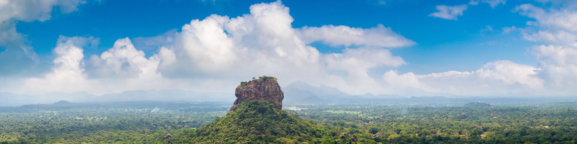 Formação rochosa imponente rodeada por floresta verde exuberante e uma paisagem extensa sob um céu azul com nuvens esparsas.