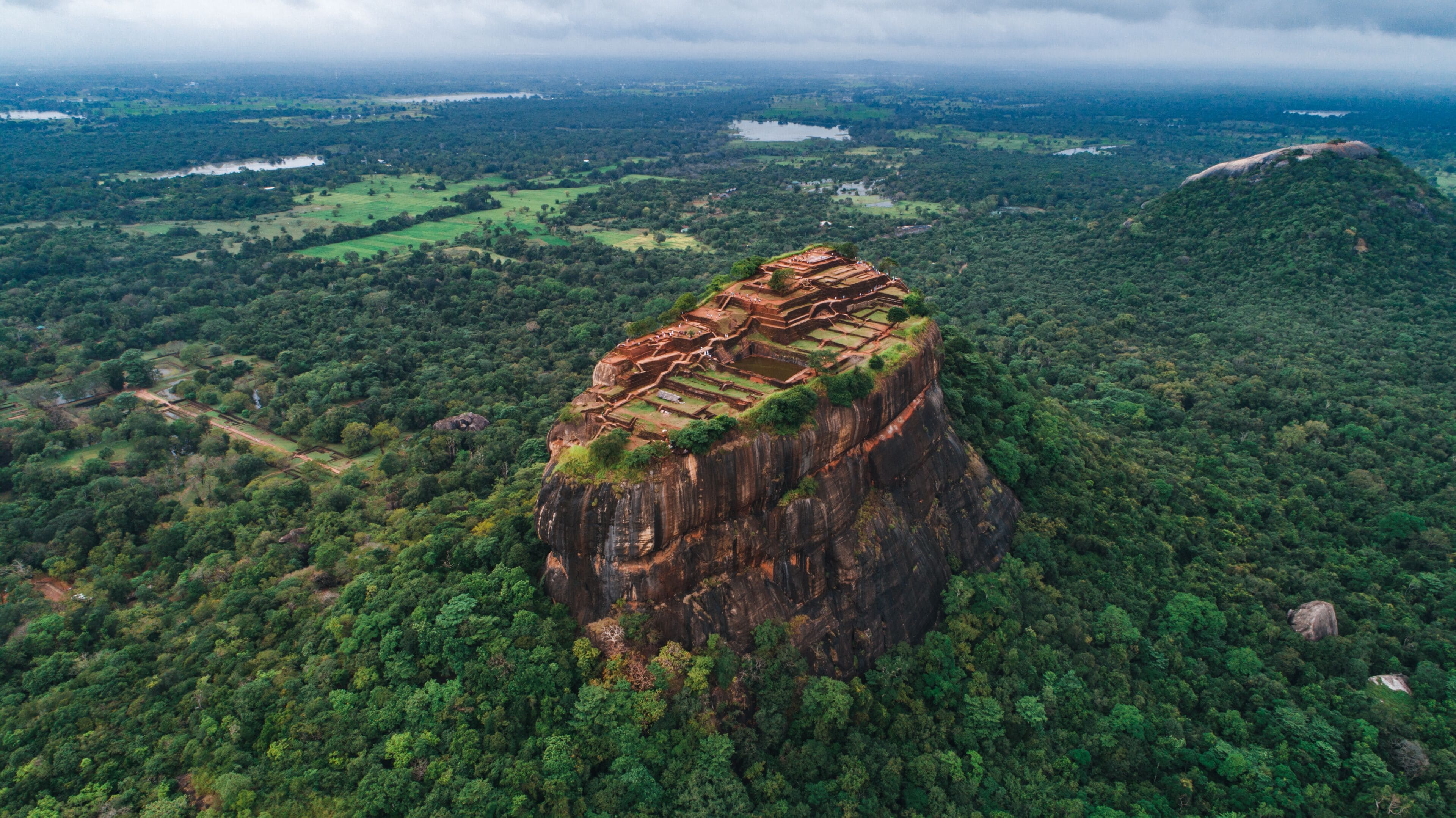 A large, rugged cliff with ancient ruins at its peak, surrounded by dense forest and a distant lake.