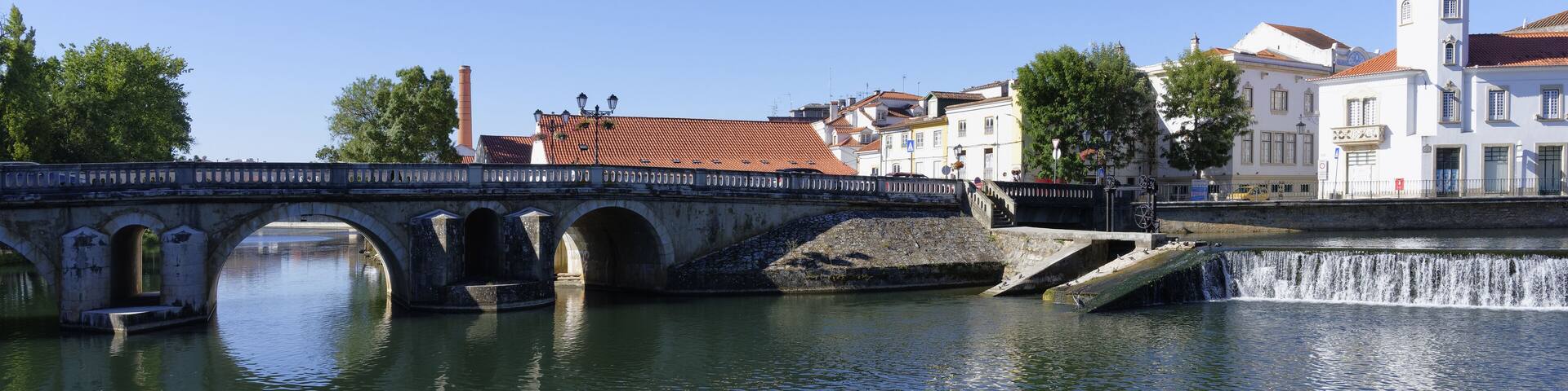 A stone bridge over a calm river, surrounded by historic buildings and a church with a red roof.