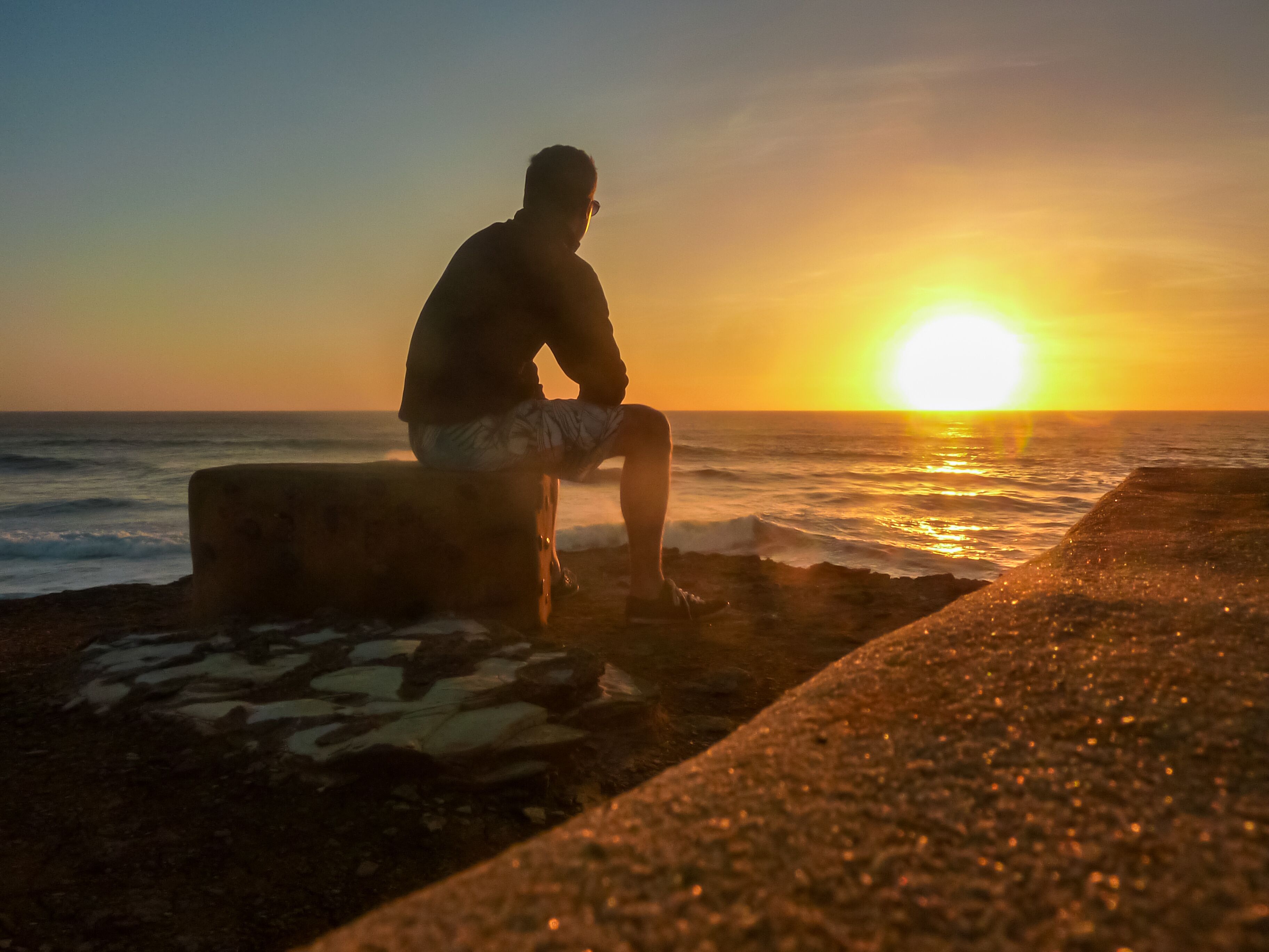 Tourist man sitting on stone bench watching romantic sunset overlooking the calm waters of the sea in Ericeira, Portugal, Europe. Looking at majestic Atlantic Ocean. Serene tranquil vacation concept