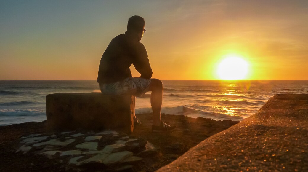 Tourist man sitting on stone bench watching romantic sunset overlooking the calm waters of the sea in Ericeira, Portugal, Europe. Looking at majestic Atlantic Ocean. Serene tranquil vacation concept