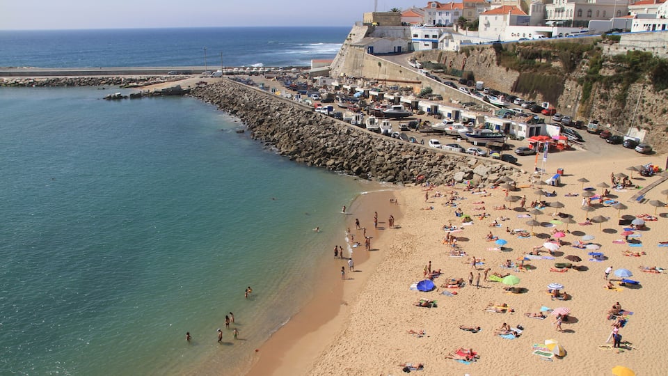 A crowded beach with people sunbathing and swimming, a rocky breakwater, and buildings in the background.