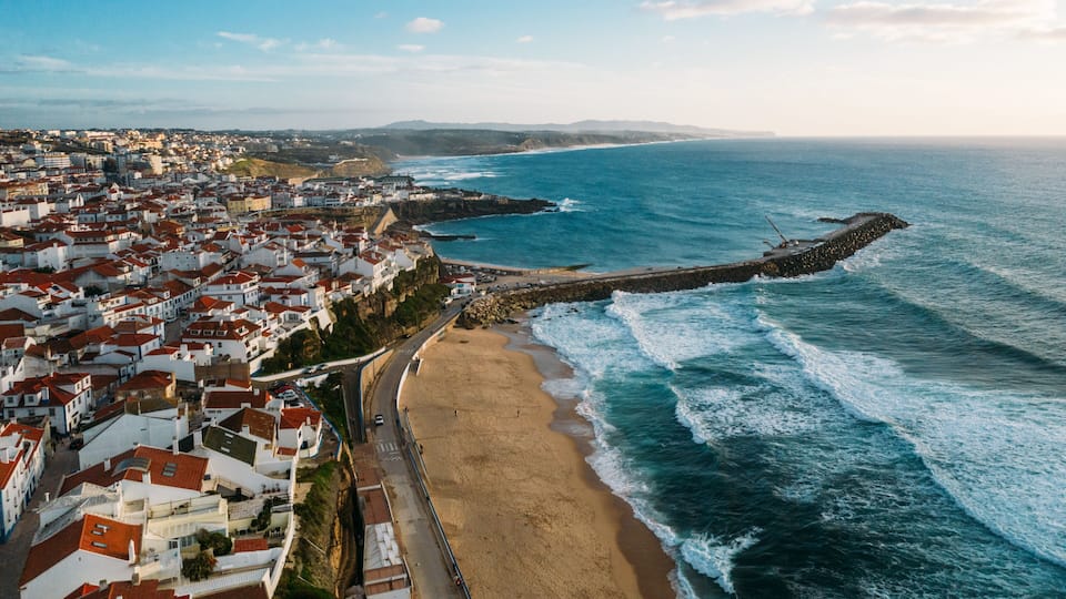 A coastal town with a beach, rocky breakwater, and red-roofed buildings.