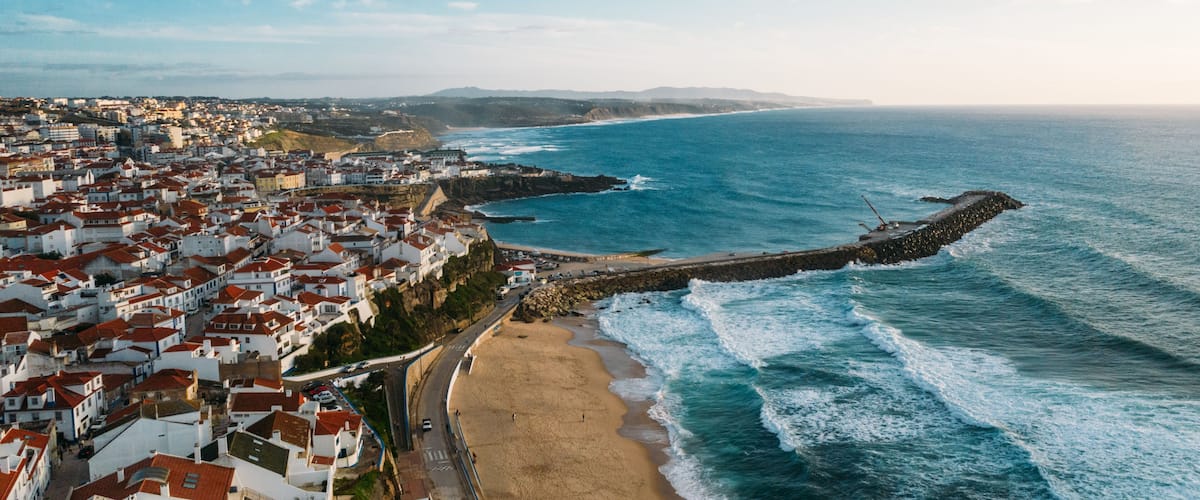 A coastal town with a beach, rocky breakwater, and red-roofed buildings.