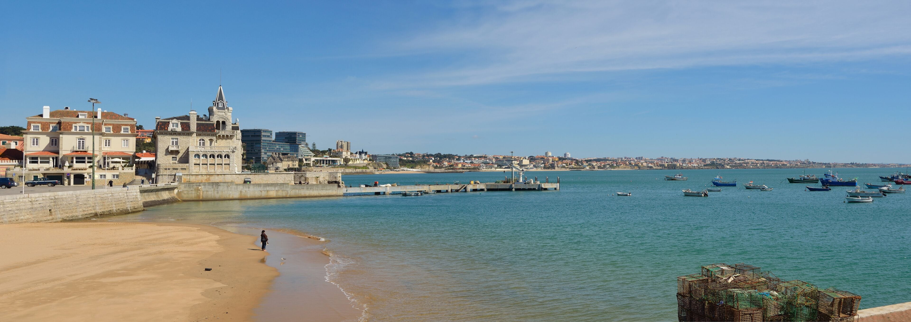 Un paisaje urbano costero con un edificio histórico, una playa de arena y un puerto con barcos.