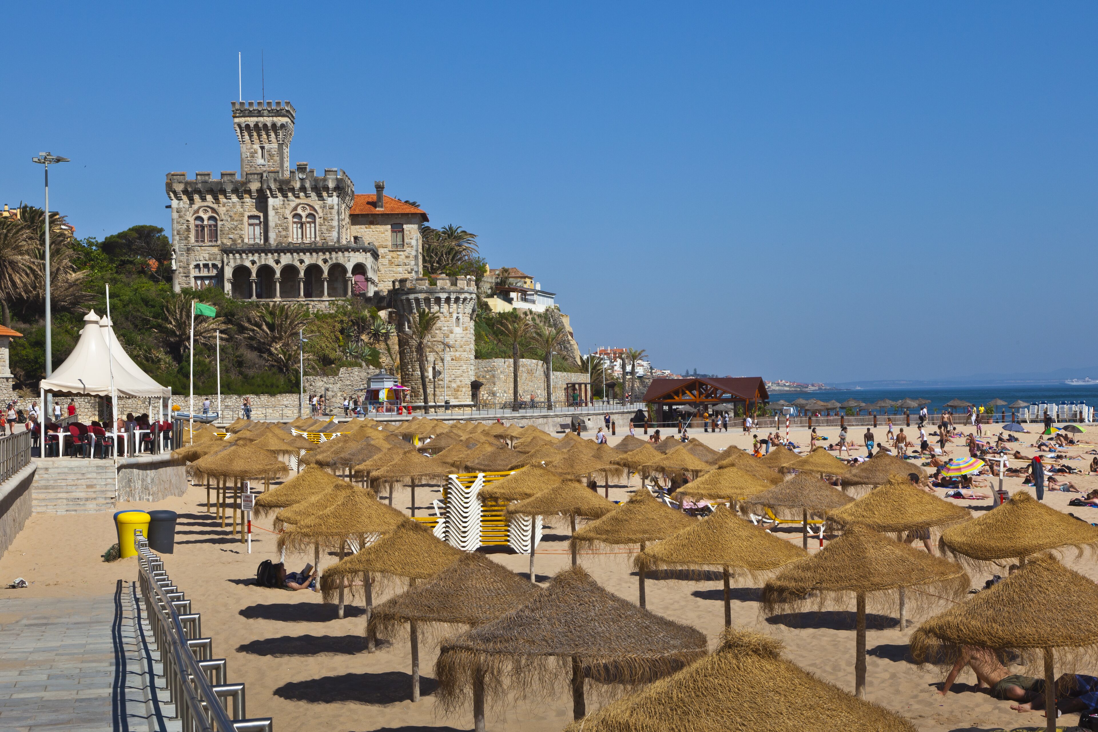 A beach with straw umbrellas and a historic castle in the background.
