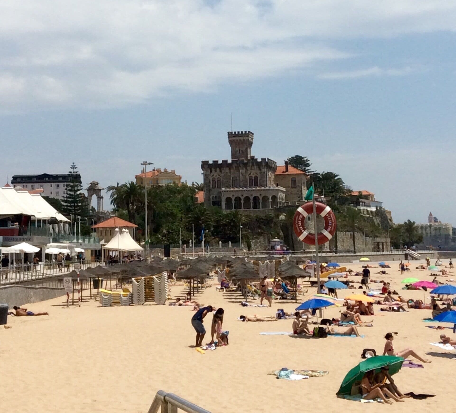 A beach with people sunbathing, umbrellas, and a castle-like building in the background.