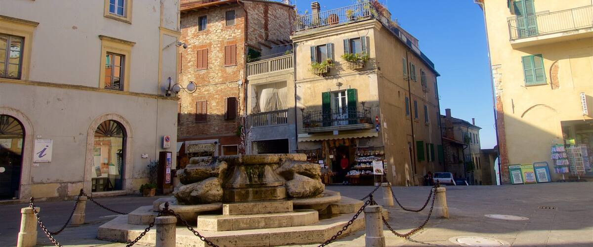A historic square with a fountain, stone steps, and traditional buildings with balconies and shutters.