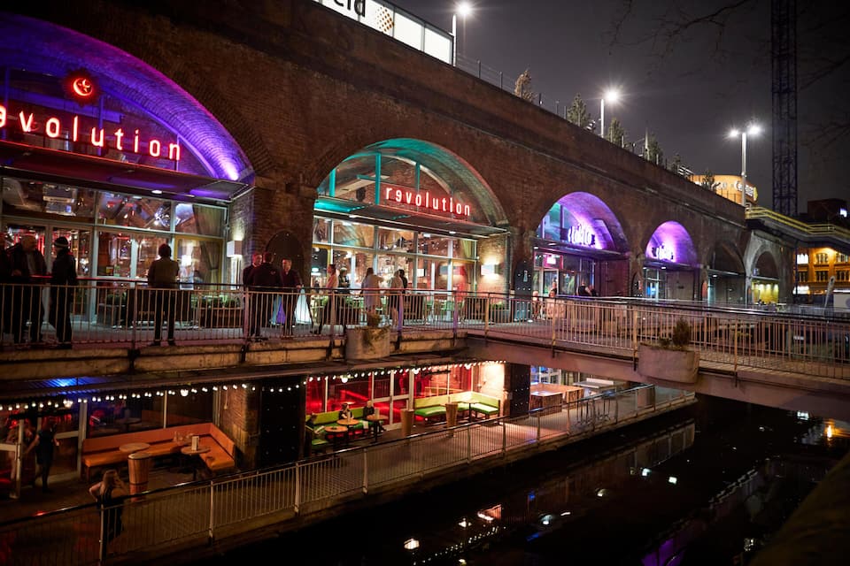 A brightly lit restaurant named 'revolution' with outdoor seating and a canal below.