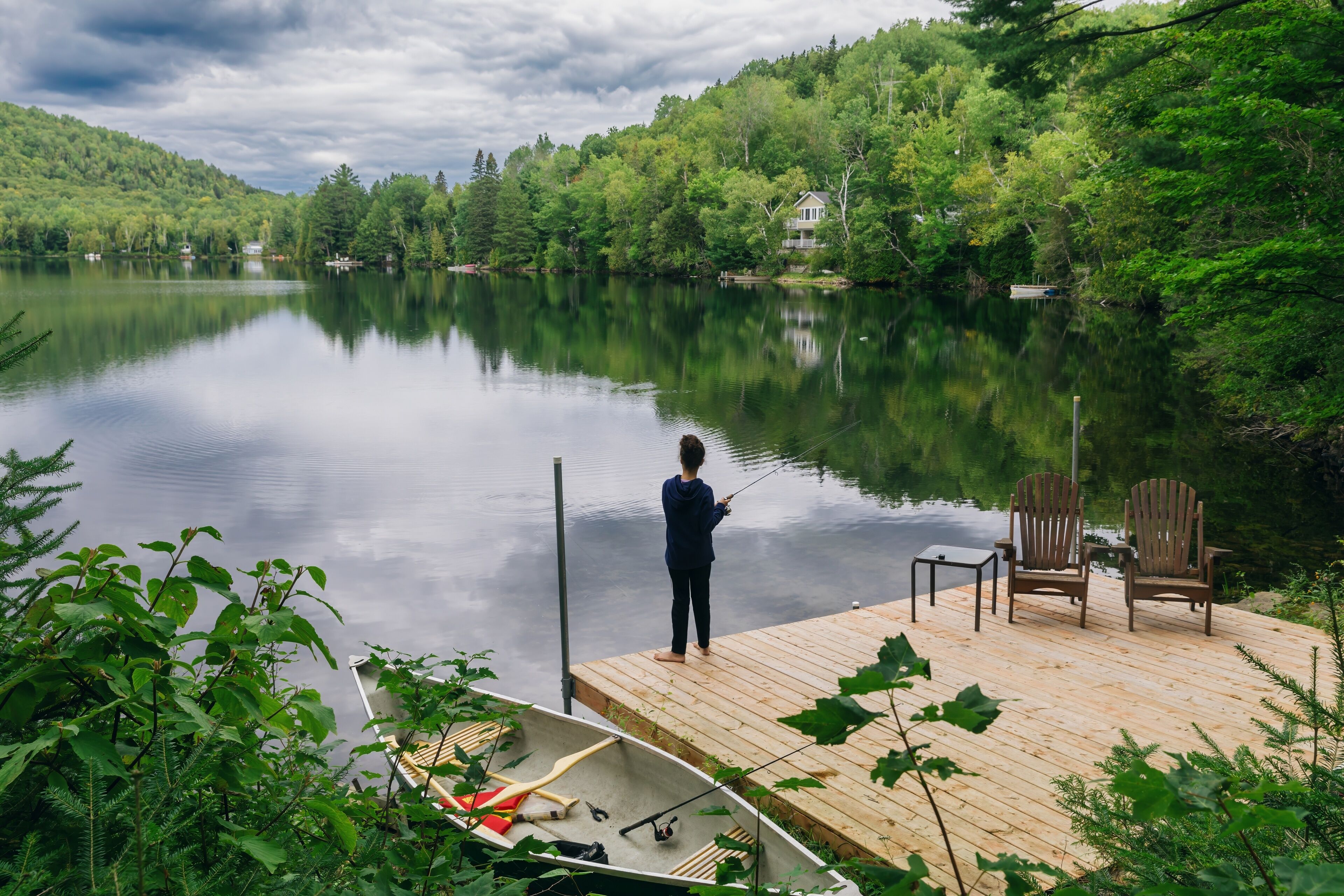 Young girl fishing off a pier in a calm tranquil lake. A canoe is moored on the shore. Lac Lambert, Saint-Alexis-des-Monts, Quebec, Canada.