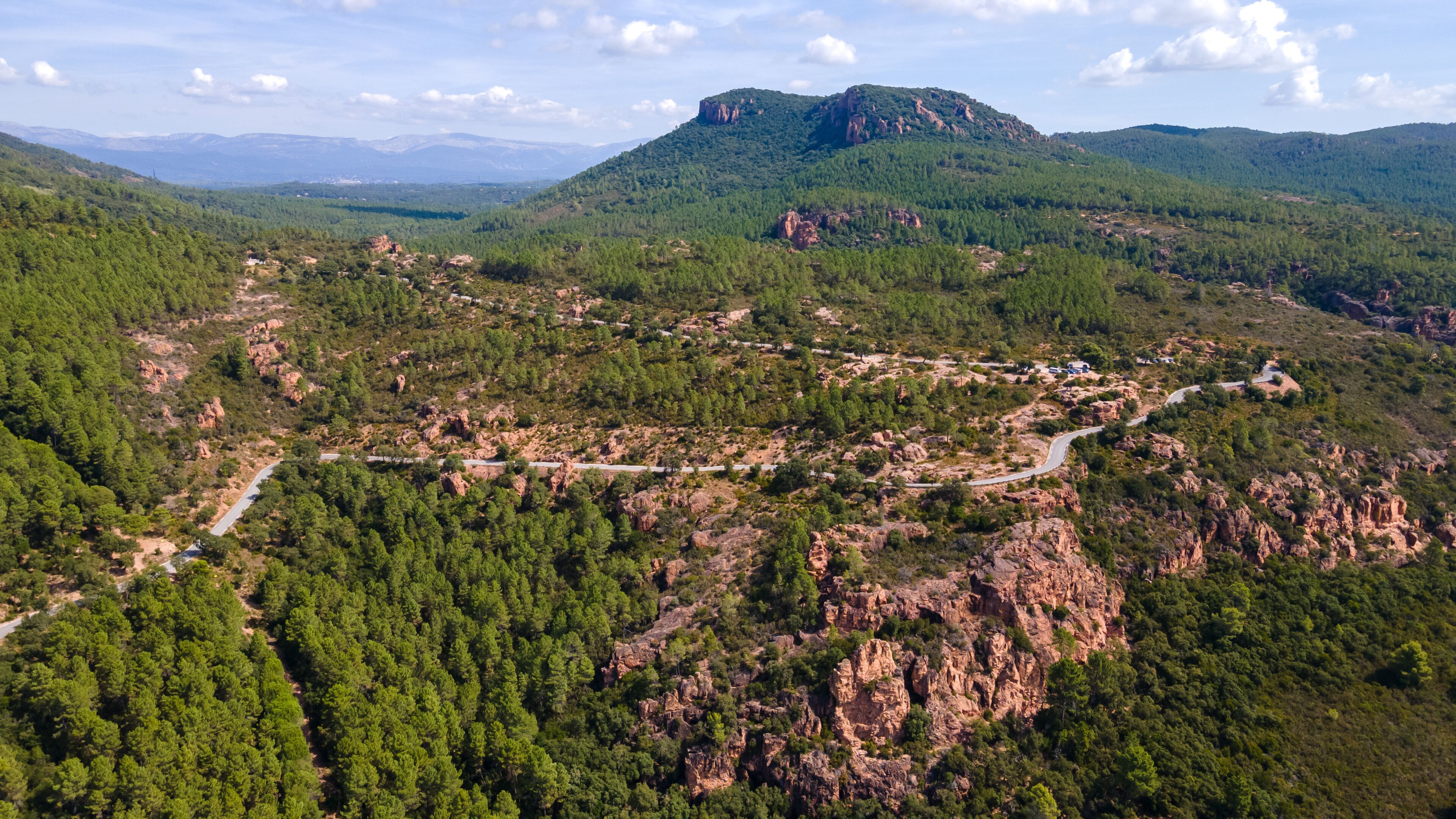 A mountainous landscape with a winding road, dense forests, and a prominent rocky peak.