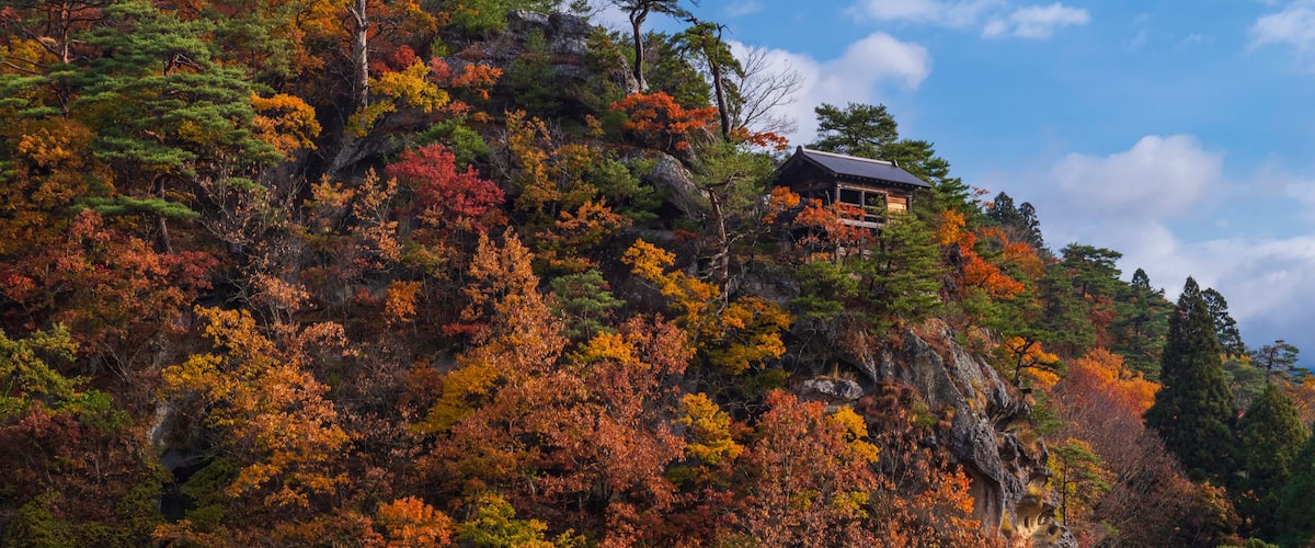 A mountain with autumn foliage and a traditional wooden building on top.