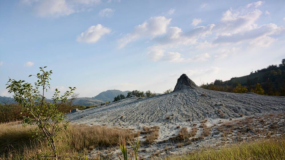 Un paysage volcanique aride, dominé par un cratère imposant, entouré d’une végétation clairsemée et bordé de collines lointaines, sous un ciel partiellement nuageux.
