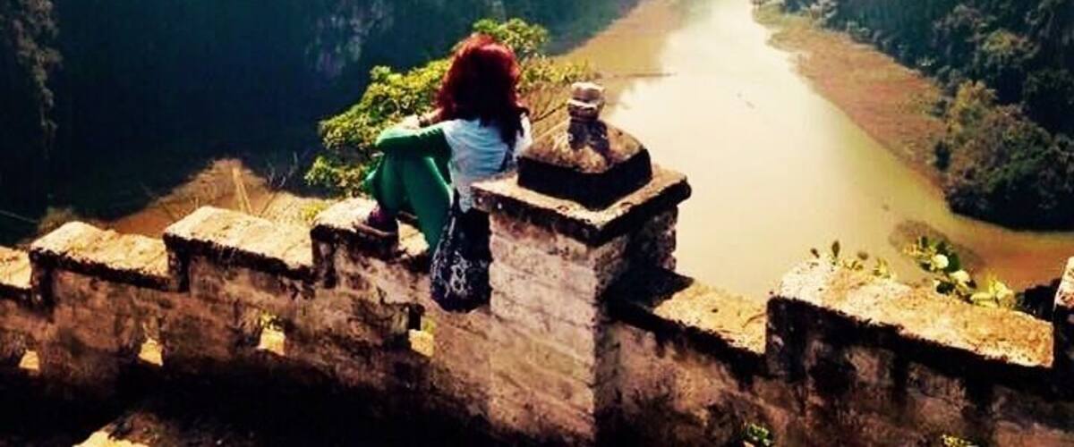A person sitting on a stone wall overlooking a river surrounded by forested hills.