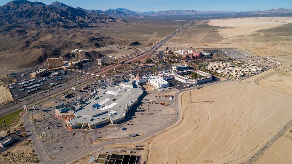 An aerial view of a desert city with a large industrial complex, a casino, and a mountain range in the background.