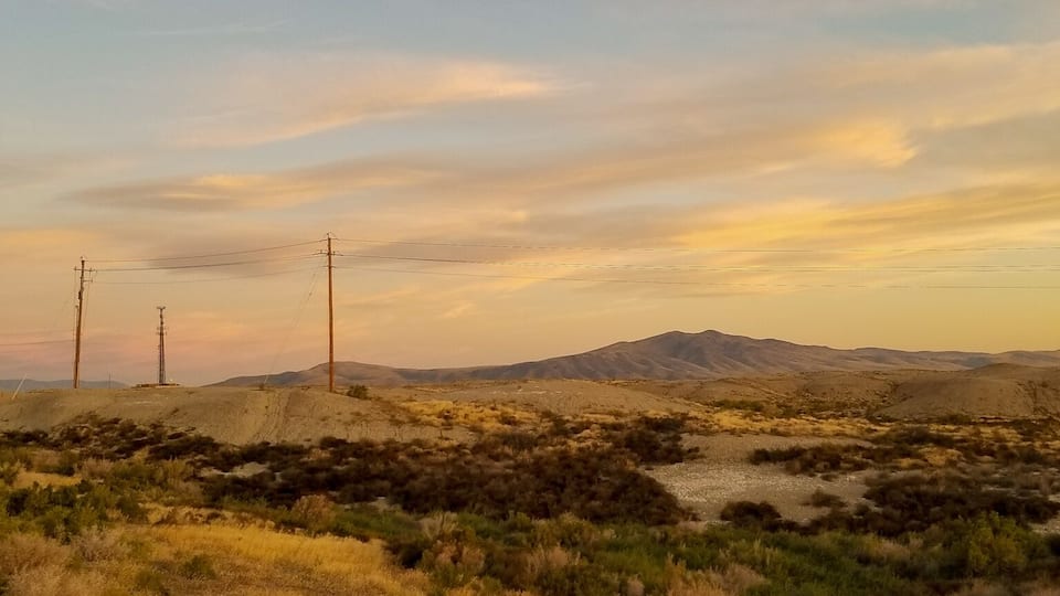 A desert landscape with sparse vegetation, power lines, and distant hills under a clear sky.